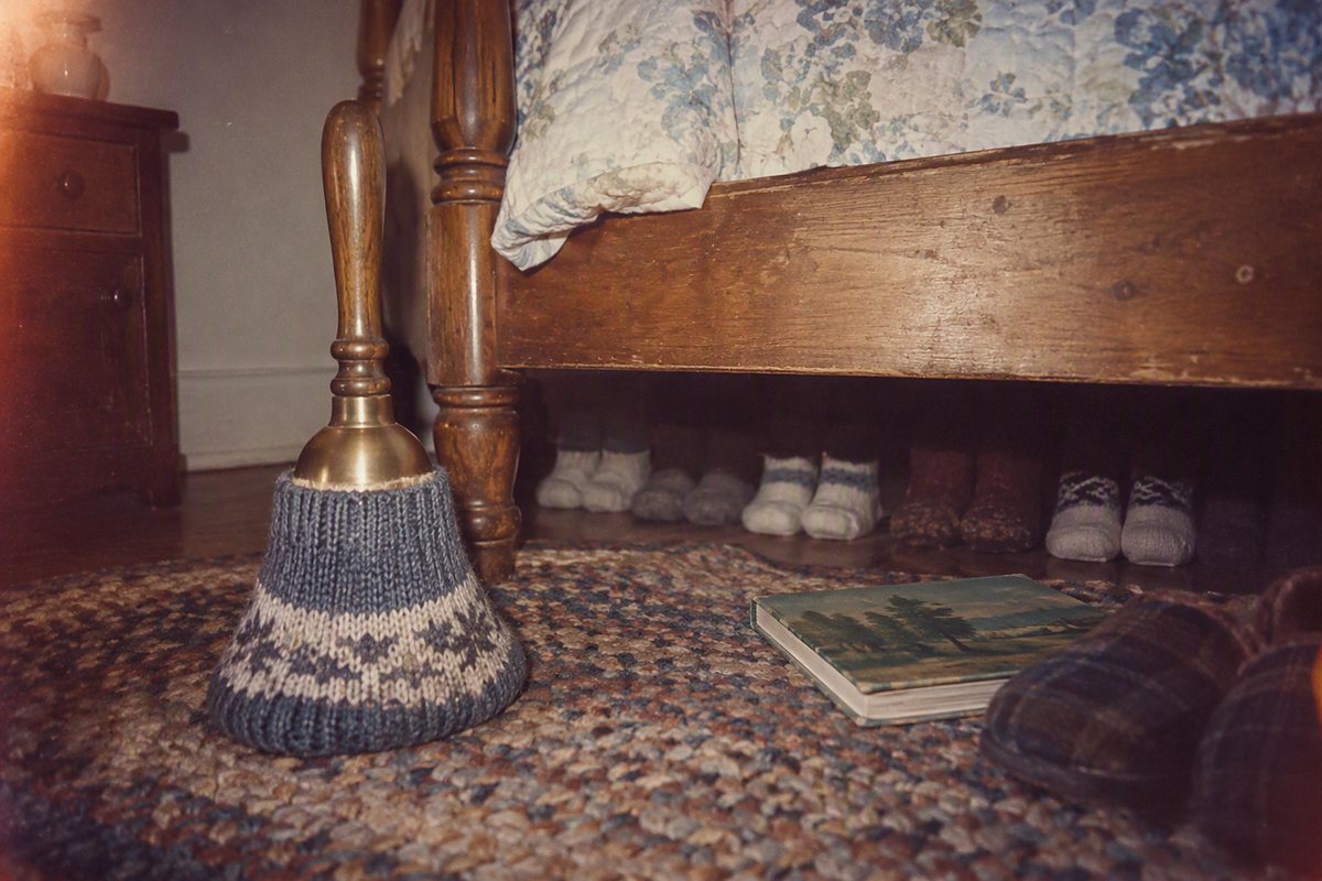A brass bell with a knitted cover stands beside a wooden bed, with shoes underneath and books nearby.